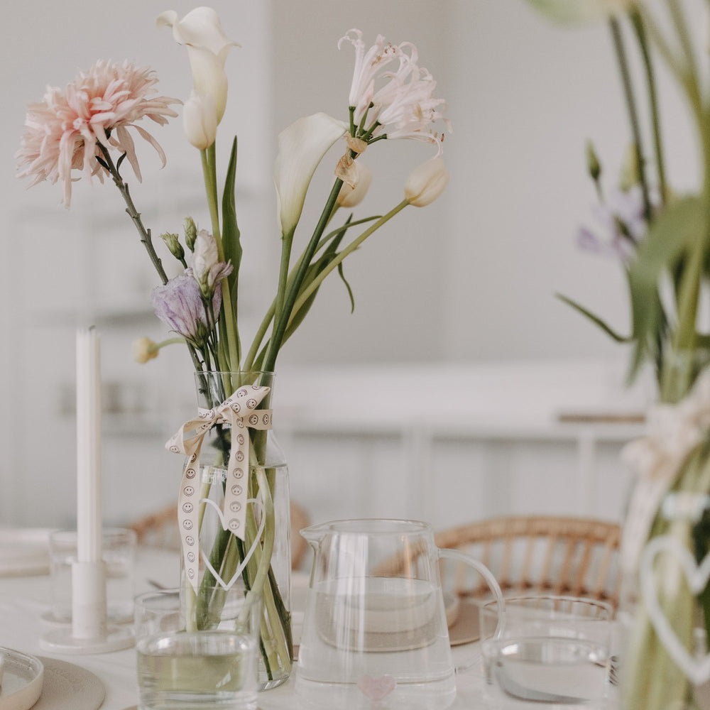 Blumenstrauß in Glasvase, dekoriert mit Smiley-Geschenkband auf festlichem Tisch.
