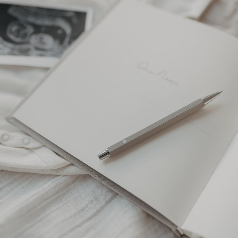 Grauer Druckbleistift auf offenem Notizbuch, umgeben von sanften Stoffen und Ultraschallbild.