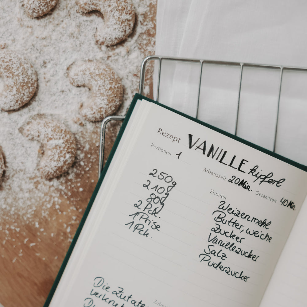 Offenes Rezeptbuch mit handgeschriebenem Vanillekipferl-Rezept neben frisch gebackenen Kipferln.