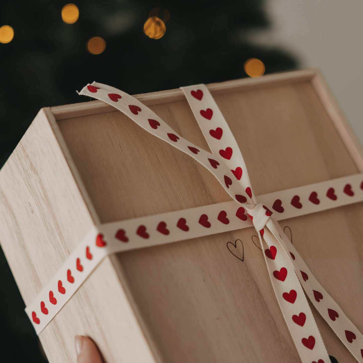 Holzbox mit beigem Geschenkband und roten Herzen vor einem Weihnachtsbaum